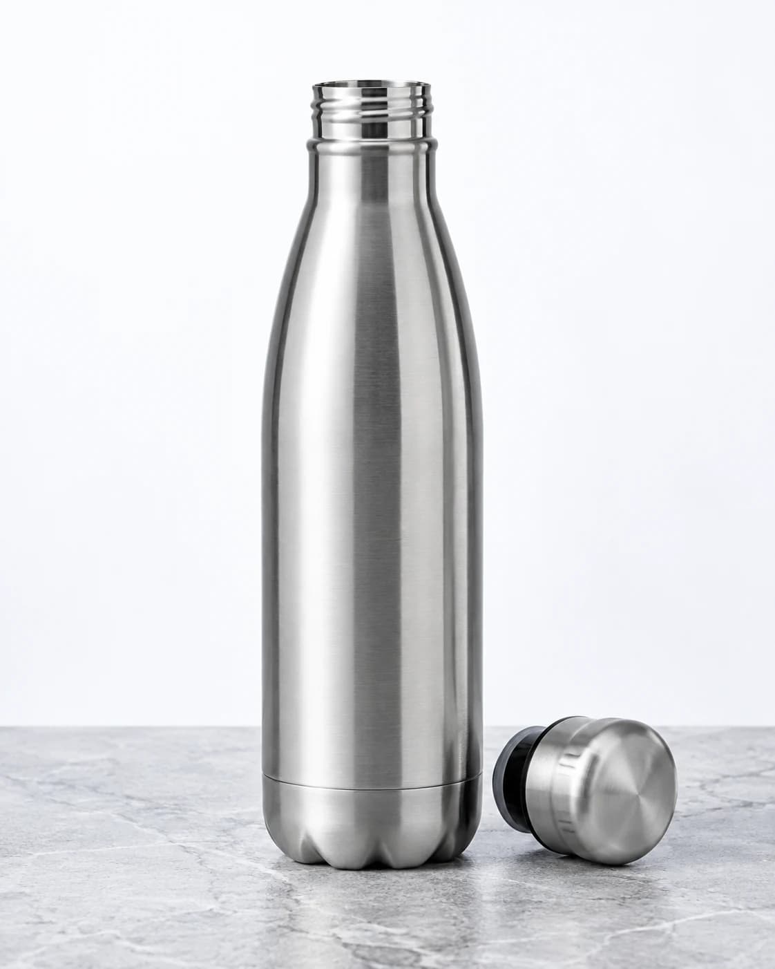 a brushed steel insulated water bottle in a light gray stone slab, a centered upright hero with its cap beside it with crisp softbox illumination